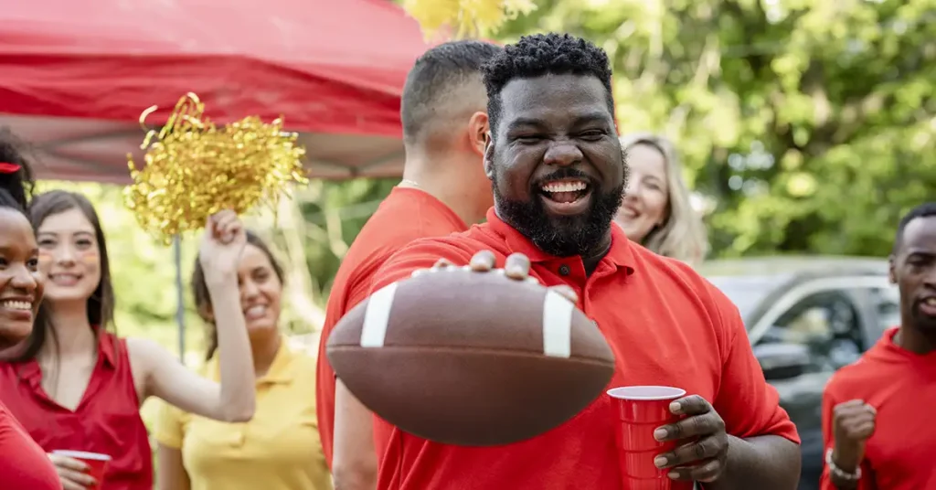 Smiling man in a red shirt holding a football at a lively tailgate party, surrounded by friends in red and yellow cheering with pom-poms and drinks.